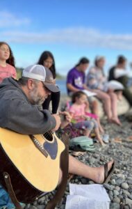 Man with goatee plays a guitar sitting on rocky beach. Several youth on a driftwood log behind him singing along.